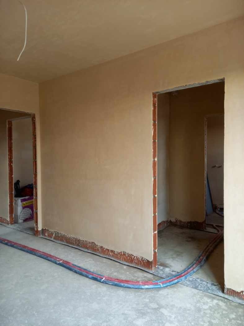 Wnętrze w trakcie prac wykończeniowych: ściany pokryte świeżo nałożoną, jasną warstwą gładzi, widoczne fragmenty cegieł przy podłodze i ościeżnicach, na podłodze leżą węże instalacyjne.