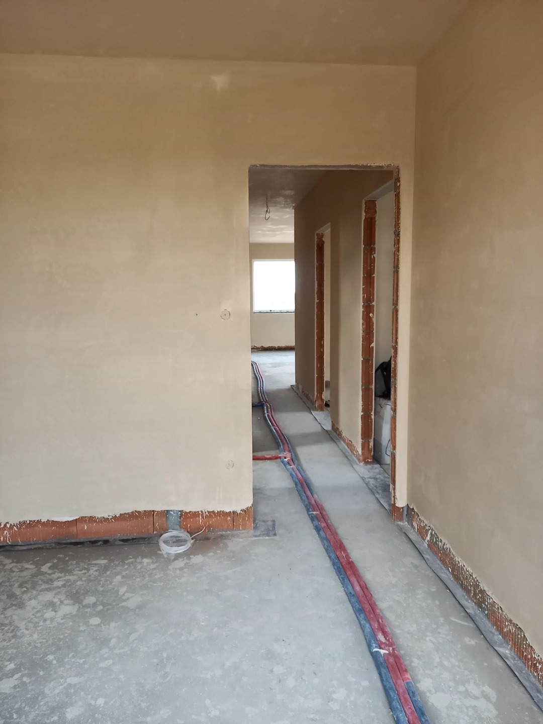 Wnętrze w trakcie tynkowania maszynowego: ściany pokryte świeżym tynkiem, widoczne instalacje w podłodze i fragmenty cegieł przy podłodze.