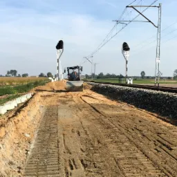 Walec drogowy ubija ziemię wzdłuż torów kolejowych; widoczne słupy trakcyjne i semafory.