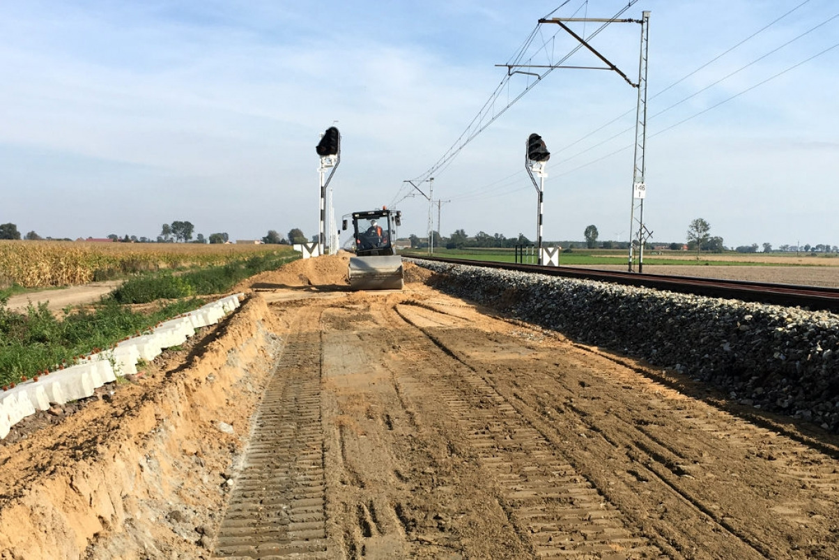 Walec drogowy ubija ziemię wzdłuż torów kolejowych; widoczne słupy trakcyjne i semafory.