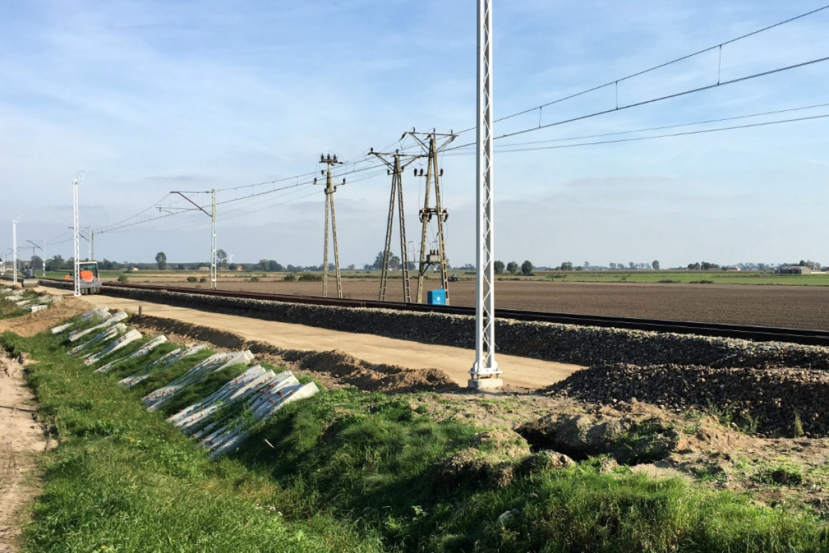 Widok na linię kolejową w trakcie budowy lub remontu, z widocznymi słupami trakcyjnymi, podsypką tłuczniową i fragmentami betonu w trawie.