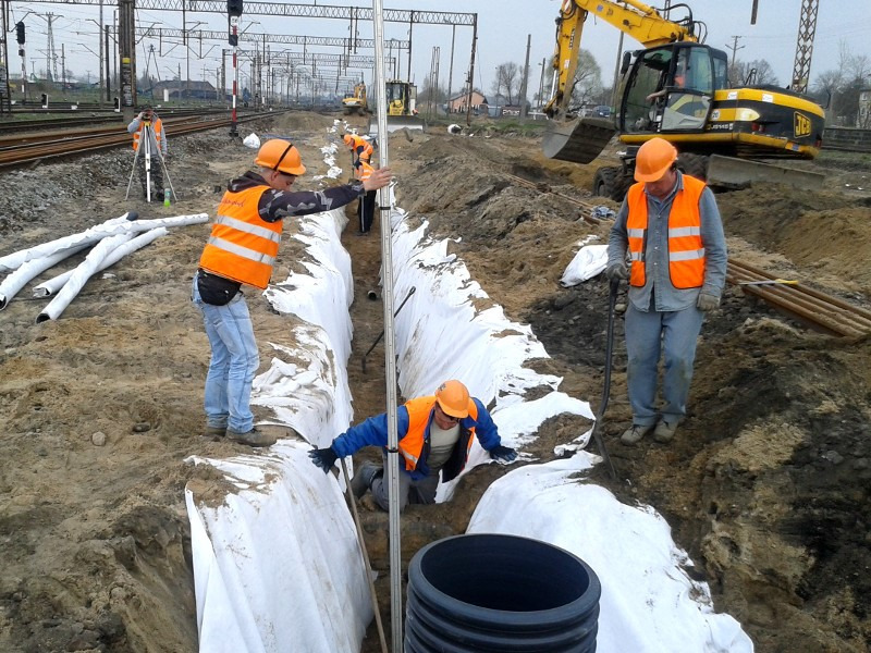 Pracownicy w pomarańczowych kamizelkach i kaskach układają rury w wykopie zabezpieczonym białą geowłókniną, w tle torowisko kolejowe i koparka JCB.