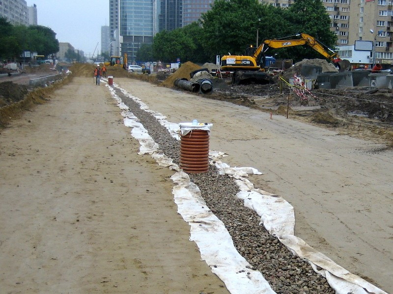 Budowa drogi: Wykop z drenażem wzdłuż przyszłej jezdni, geowłóknina, żwir, rury drenarskie, pomarańczowa studzienka kanalizacyjna, koparka JCB w tle, robotnicy w odblaskowych kamizelkach.