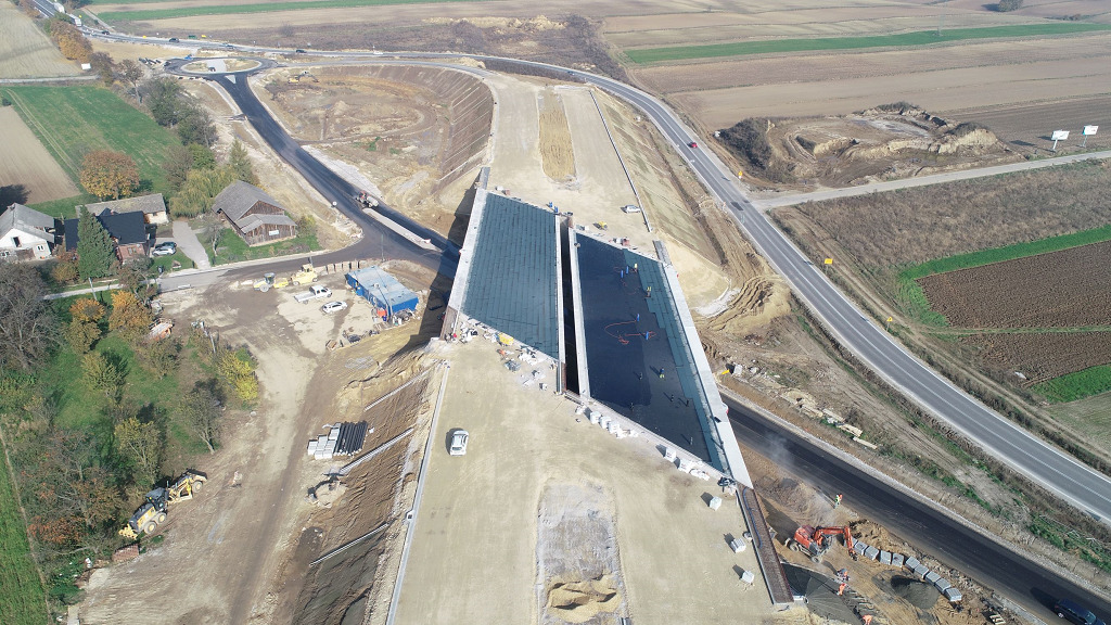 Widok z lotu ptaka na plac budowy z dwiema ukośnymi konstrukcjami, prawdopodobnie częścią węzła drogowego, z pracującymi robotnikami i sprzętem budowlanym, otoczony polami i drogami.