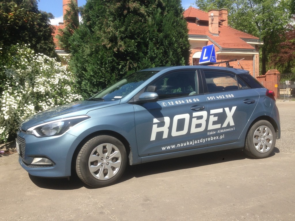 Niebieski samochód z oznaczeniem 'L' na dachu, logo 'ROBEX' i numerami kontaktowymi na boku, zaparkowany na tle budynku z czerwonym dachem i zielenią.