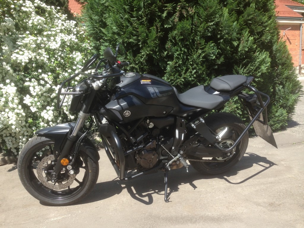 Czarny motocykl Yamaha MT-07 z dodatkowymi orurowaniami ochronnymi i stelażem pod sakwy, zaparkowany na betonowym podłożu, na tle kwitnącego krzewu i żywopłotu.