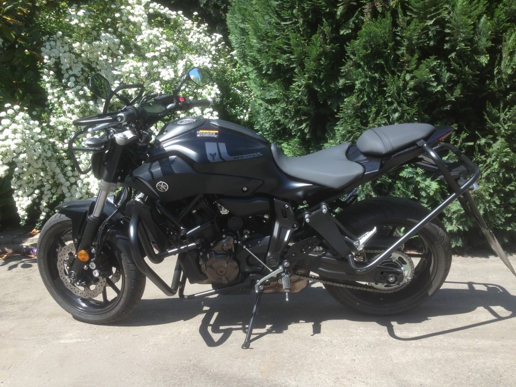 Czarny motocykl Yamaha MT-07 na tle kwitnącego krzewu i żywopłotu.
