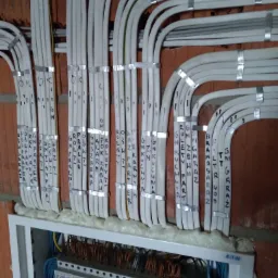 Rozdzielnica elektryczna z licznymi przewodami oznaczonymi markerem, przymocowanymi metalowymi opaskami do ściany z czerwonej cegły. Widoczna piana montażowa uszczelniająca przestrzeń wokół przewodów.