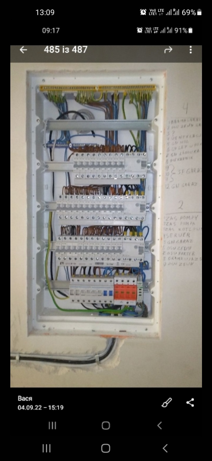 Otwarta skrzynka rozdzielcza z widocznymi przewodami, bezpiecznikami i listwami zaciskowymi, zamontowana na ścianie z notatkami odręcznymi.