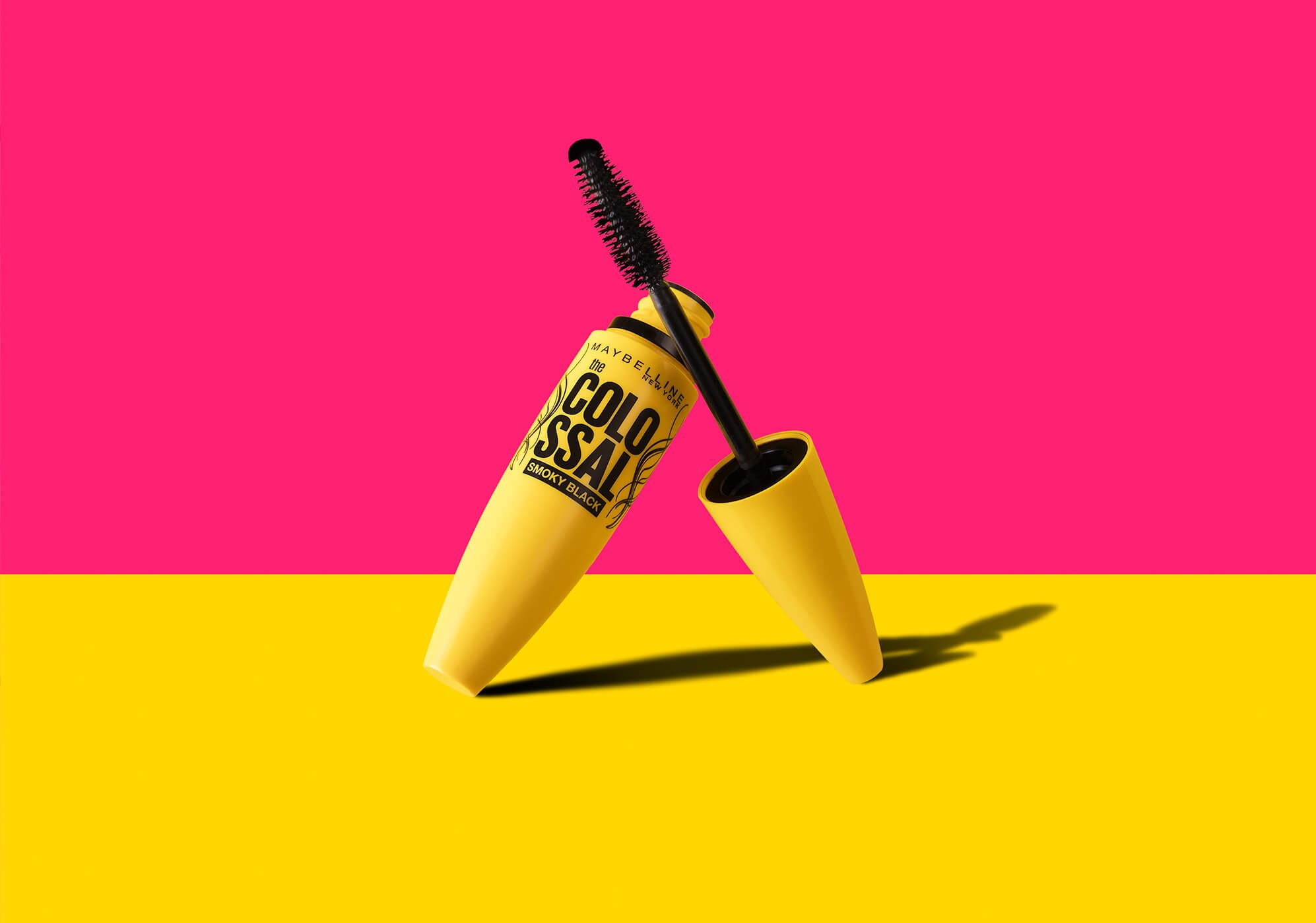 Żółta maskara Maybelline Colossal Smoky Black, z otwartą tubką i szczoteczką, na dwukolorowym tle: różowym i żółtym.