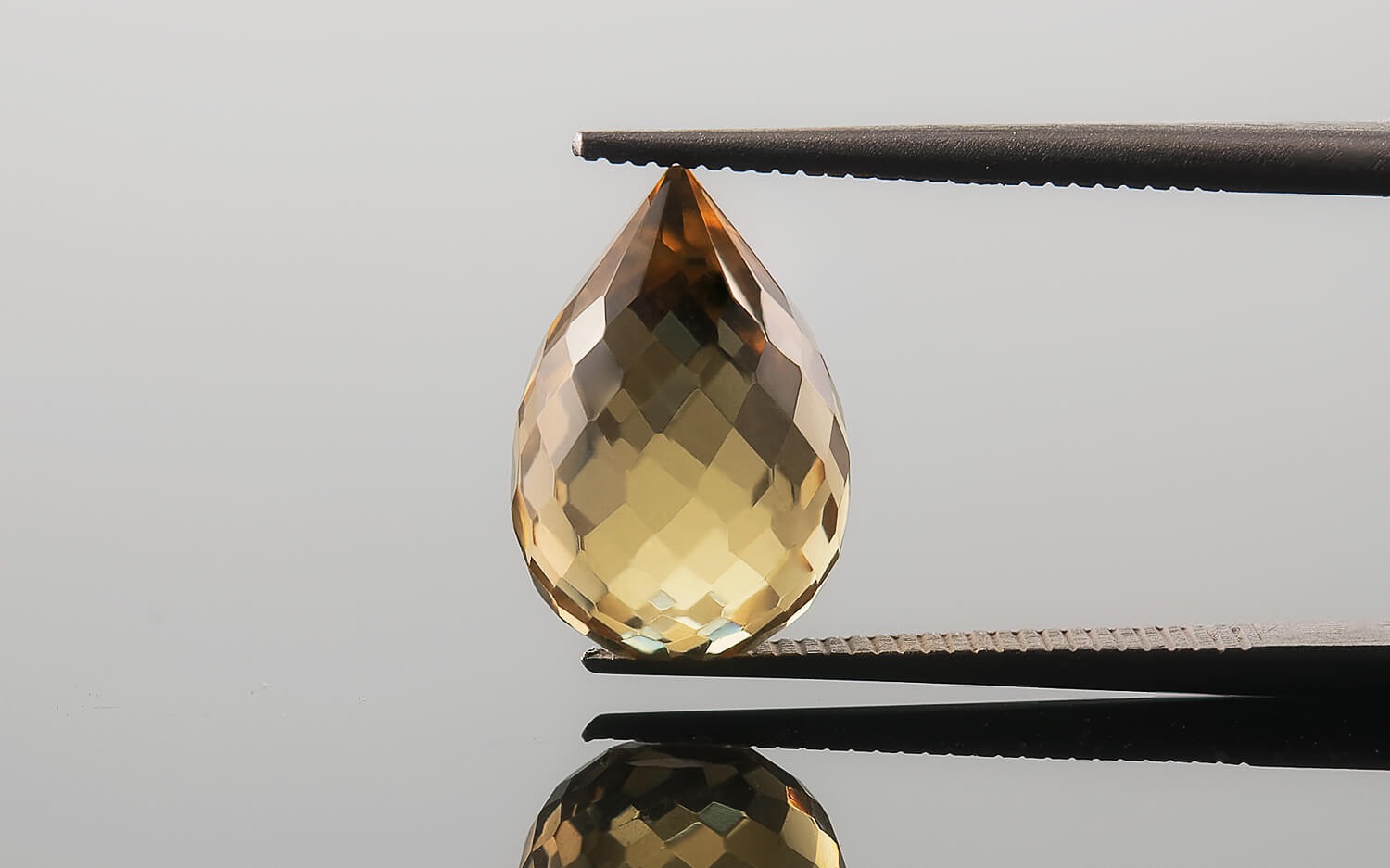 Faceted citrine gemstone, teardrop-shaped, held by tweezers against a reflective grey background, showing its internal facets and golden hues.