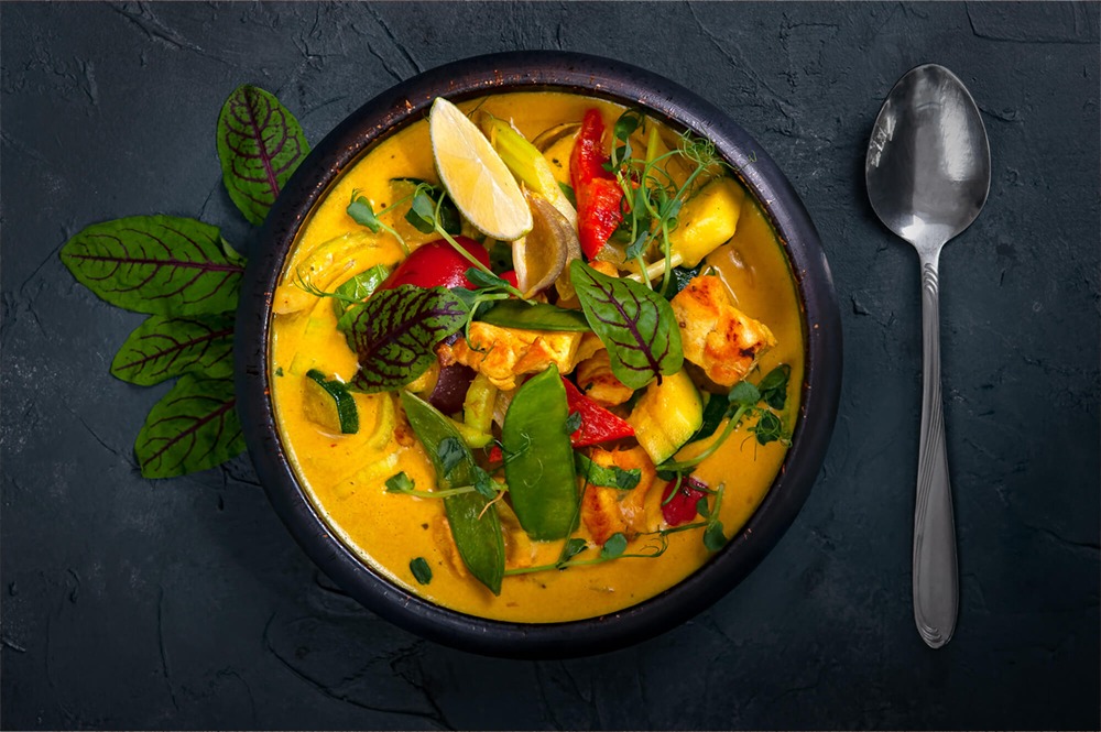 Apetyczne, żółte curry z kurczakiem, warzywami i limonką w czarnej misce, sfotografowane z góry we Wrocławiu, obok leży srebrna łyżka na ciemnym tle.