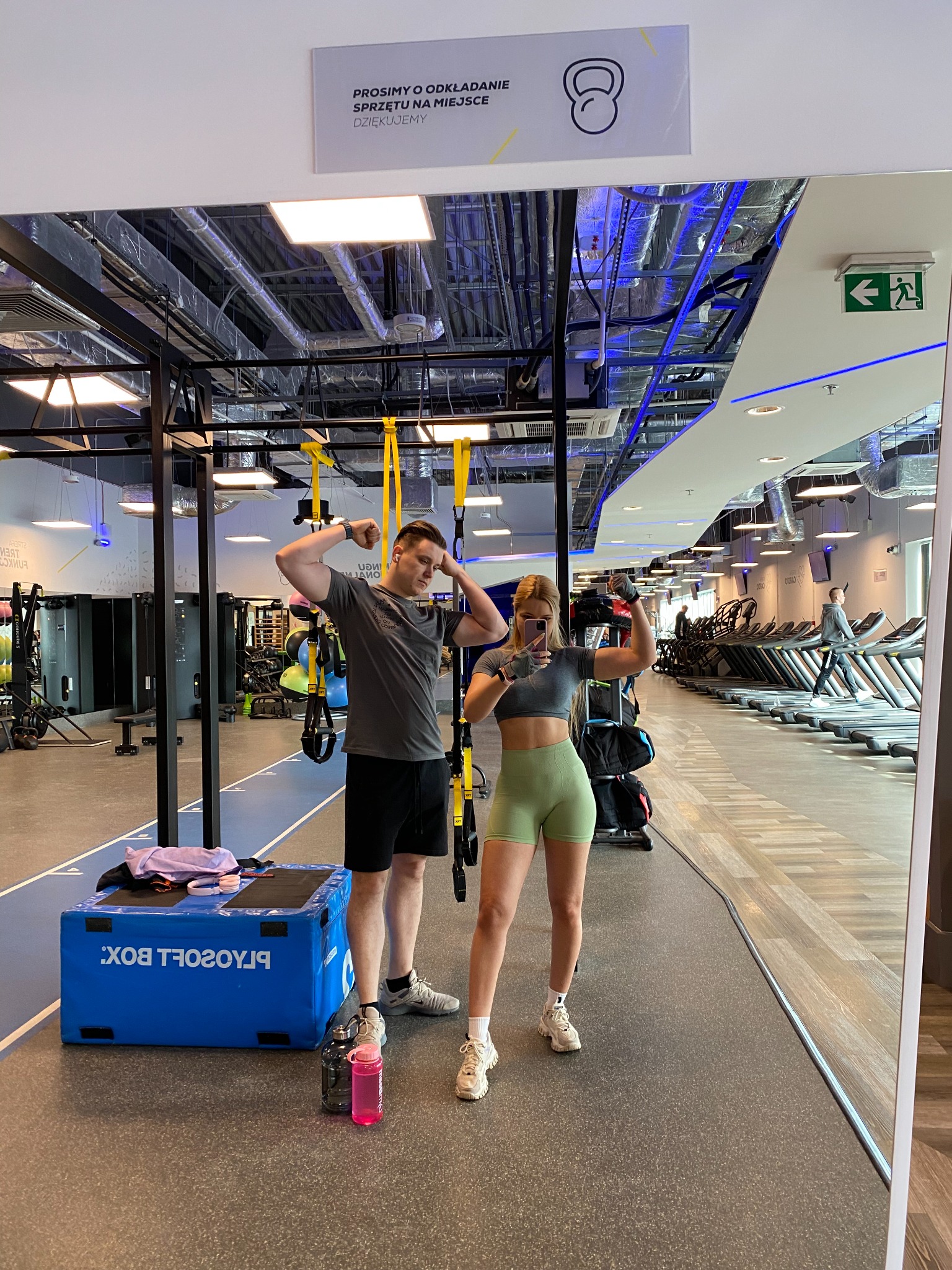 Para w sportowych strojach prezentuje bicepsy przed lustrem w nowoczesnej siłowni z widocznym sprzętem do ćwiczeń i bieżniami w tle.