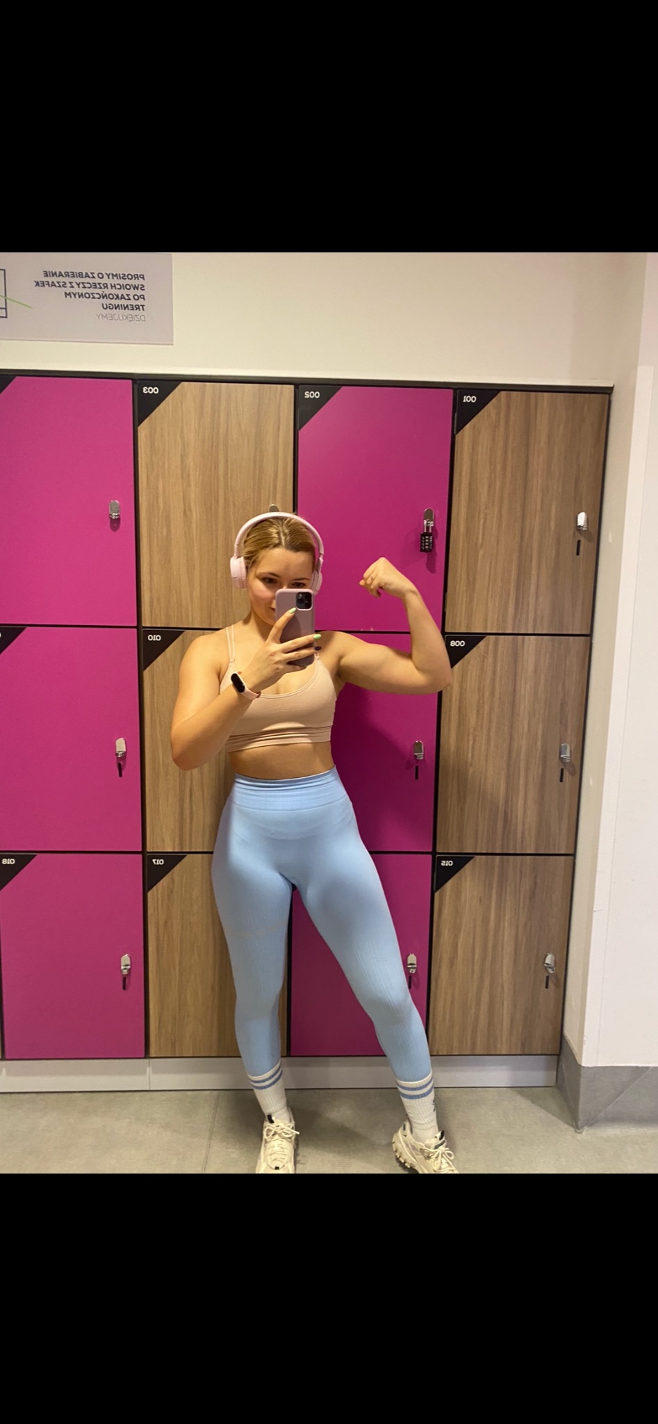 Blondynka w sportowym stroju, z białymi słuchawkami na uszach, robi selfie w siłowni, prezentując biceps na tle szafek z numerami.