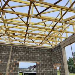 Serhii Popov - Konstrukcja drewniana więźby dachowej z metalowymi łącznikami, widok od wewnątrz budynku w trakcie budowy, ściany z szarych bloczków betonowych, pracownik na zewnątrz, błękitne niebo w tle.