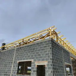 Surowy stan budynku z betonu komórkowego i drewnianą konstrukcją dachu w trakcie budowy, widoczny komin i częściowo pochmurne niebo.