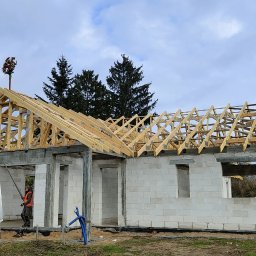 Serhii Popov - Budowa domu jednorodzinnego w stanie surowym otwartym, widoczna drewniana konstrukcja dachu z wieńcem dożynkowym na szczycie, pracownik na drabinie oparty o samochód dostawczy firmy budowlanej.