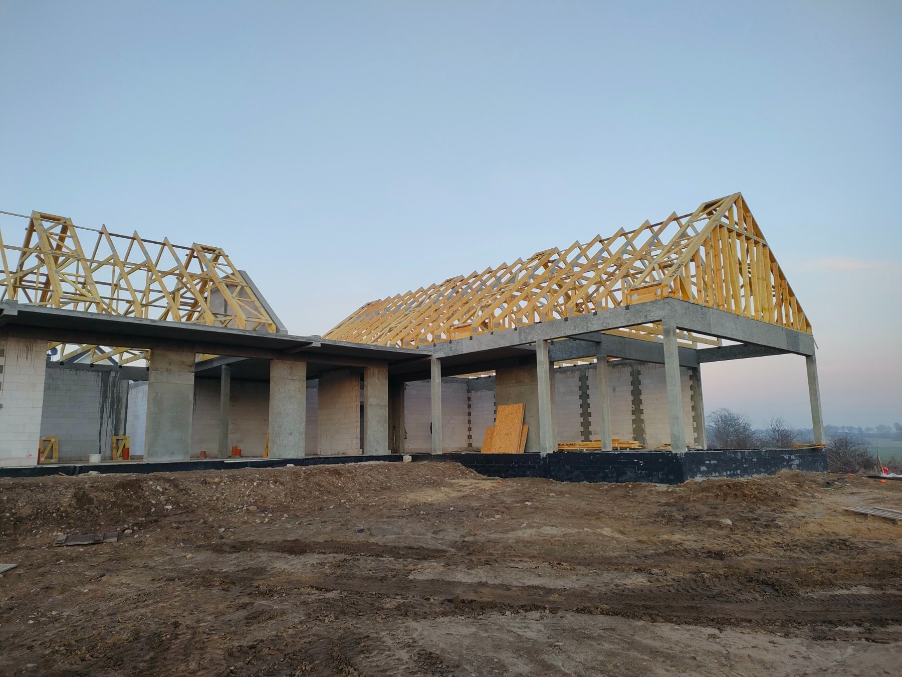 Surowa konstrukcja domu w trakcie budowy, widoczna drewniana rama dachu i betonowe ściany, ziemisty teren wokół.