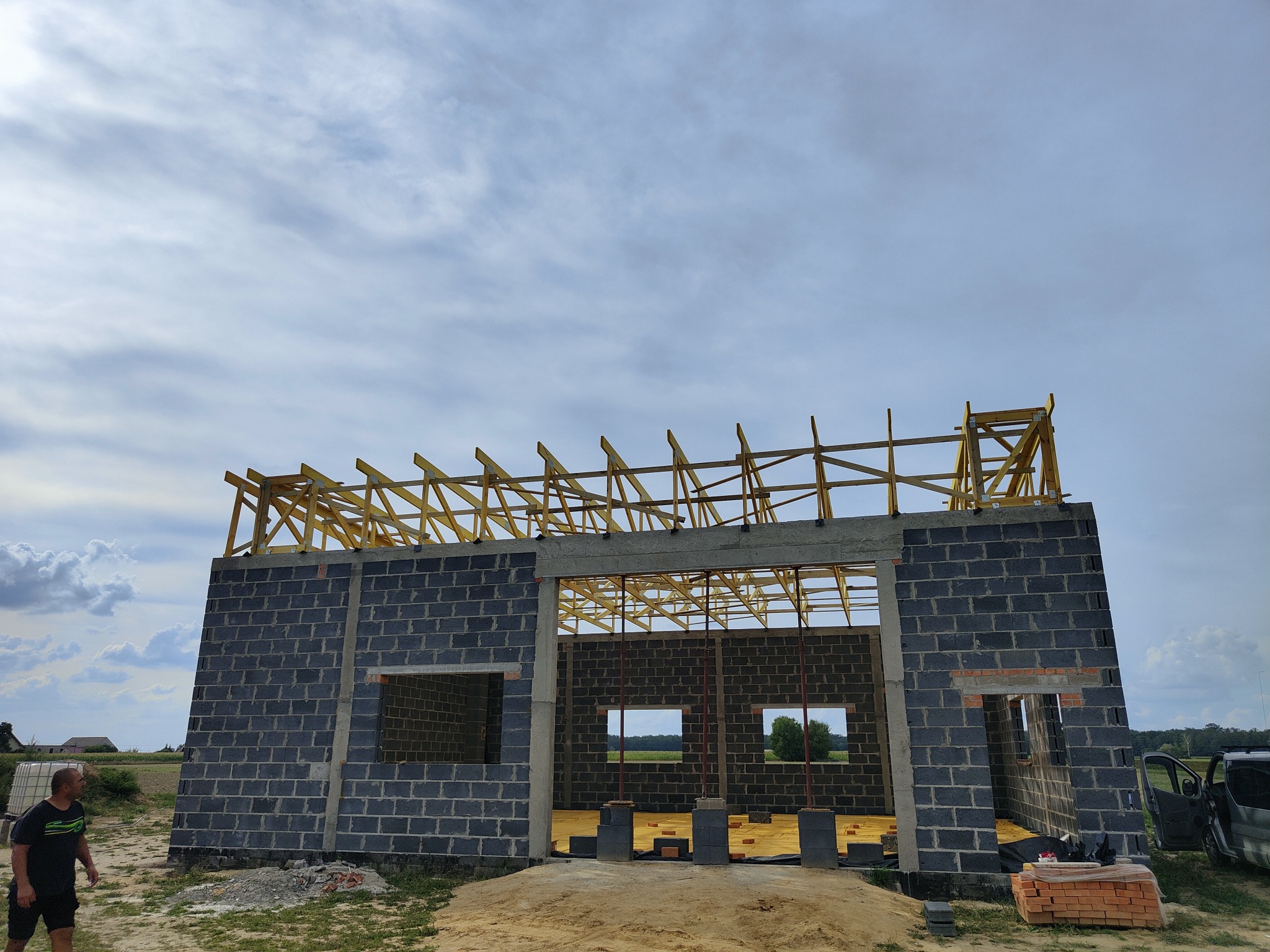 Wznoszenie ścian z bloczków i montaż drewnianej więźby dachowej na budynku w stanie surowym otwartym, widok na konstrukcję pod błękitnym niebem z chmurami.
