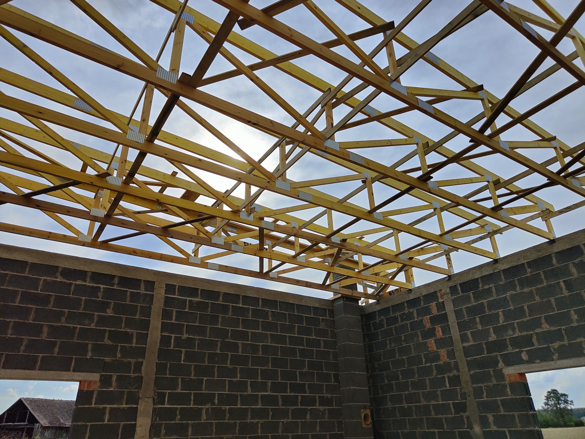 Konstrukcja drewniana więźby dachowej w trakcie montażu na murowanych ścianach budynku, widoczne metalowe łączniki i fragment nieba w tle.