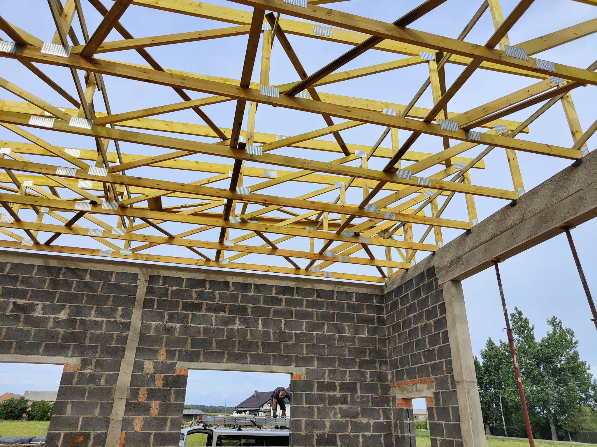 Konstrukcja drewniana więźby dachowej z metalowymi łącznikami, widok od wewnątrz budynku w trakcie budowy, ściany z szarych bloczków betonowych, pracownik na zewnątrz, błękitne niebo w tle.