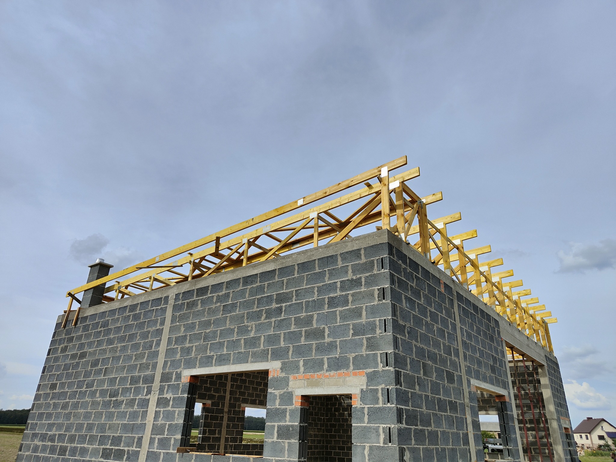 Surowy stan budynku z betonu komórkowego i drewnianą konstrukcją dachu w trakcie budowy, widoczny komin i częściowo pochmurne niebo.
