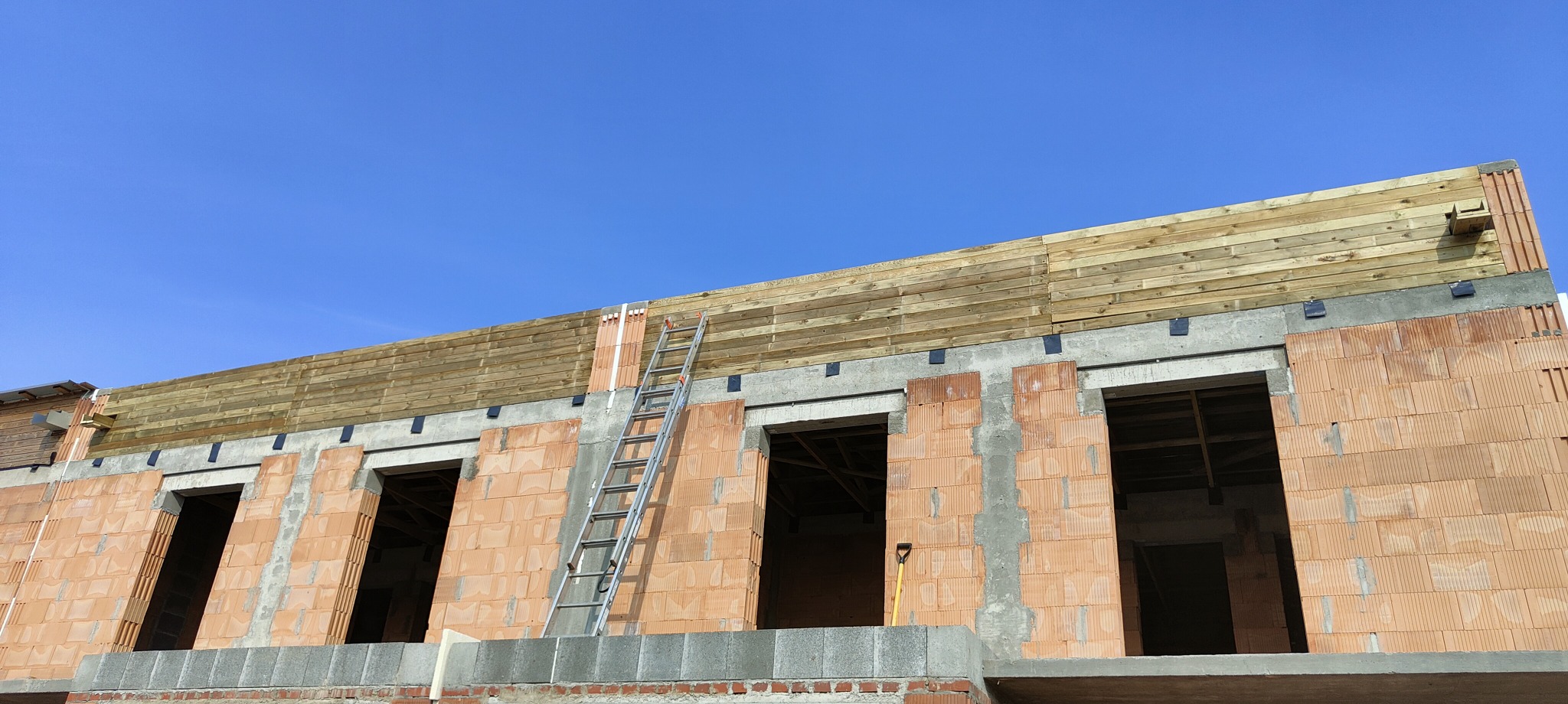 Wznoszona więźba dachowa z drewnianych belek, widoczna drabina oparta o ścianę budynku z czerwonej cegły, niebieskie niebo w tle.