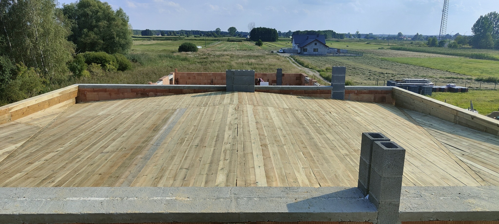 Wstępny etap budowy dachu z drewnianą konstrukcją, widoczne kominy z pustaków betonowych i ceglane ściany w tle krajobraz polny z domem.