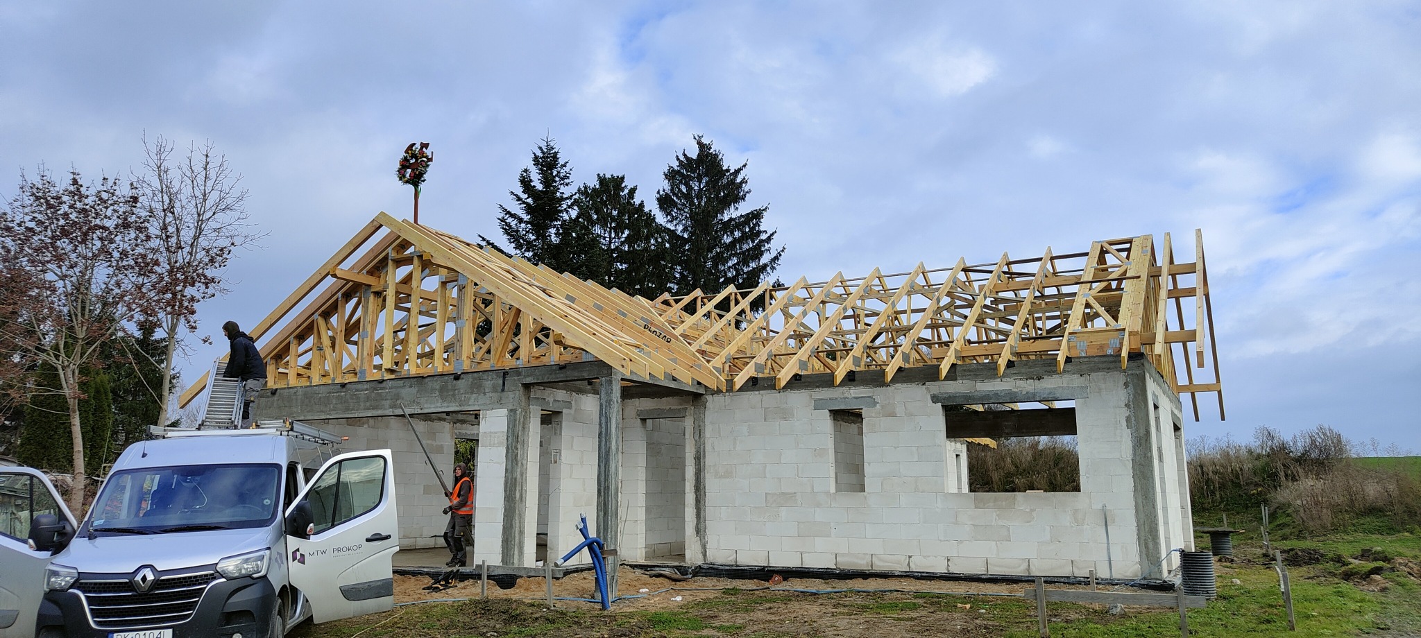 Budowa domu jednorodzinnego w stanie surowym otwartym, widoczna drewniana konstrukcja dachu z wieńcem dożynkowym na szczycie, pracownik na drabinie oparty o samochód dostawczy firmy budowlanej.