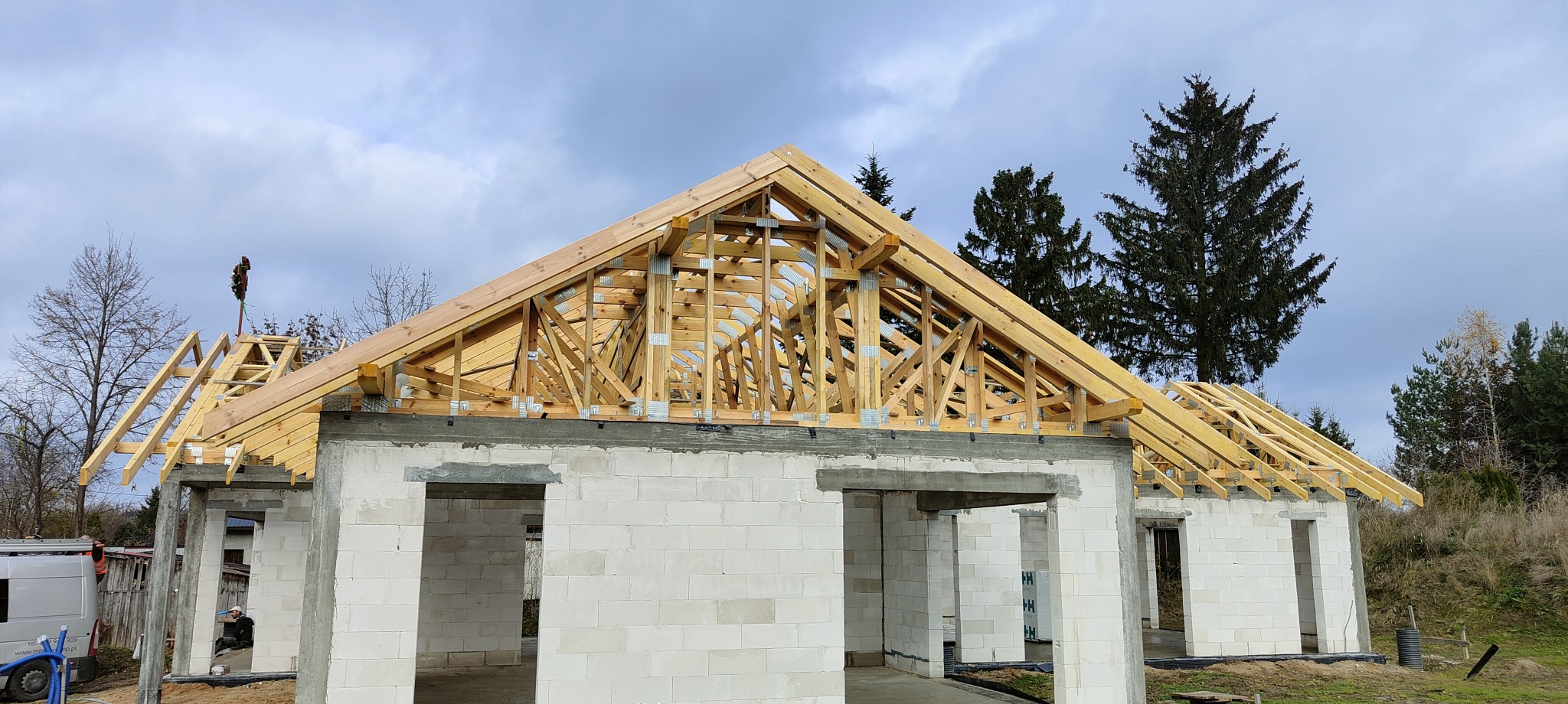 Surowa konstrukcja dachu z jasnego drewna na budynku z białego betonu komórkowego, widoczny wieniec dachowy i drzewo na szczycie dachu w trakcie budowy.