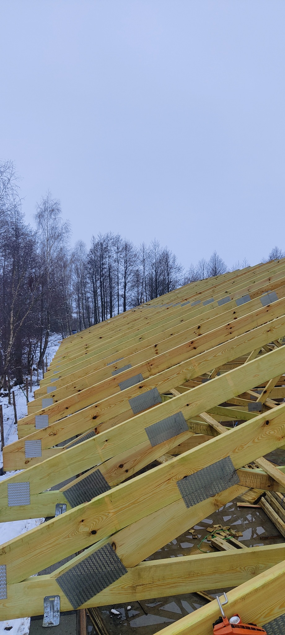 W trakcie budowy drewnianej więźby dachowej, widoczne elementy konstrukcyjne połączone metalowymi płytkami, w tle zalesiony teren w zimowej scenerii.