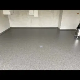 Szara, gładka podłoga w pomieszczeniu z szarymi ścianami i widocznym odpływem podłogowym oraz odkurzaczem przemysłowym w rogu.