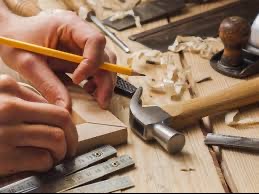 Ręce stolarza rysują ołówkiem linię na drewnianym elemencie, obok leżą narzędzia: młotek, dłuto, miarka kątowa i wióry na desce.