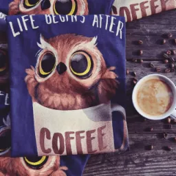 Koszula z nadrukiem sowy w filiżance kawy, leżąca na drewnianym stole obok filiżanki kawy i ziaren kawy.
