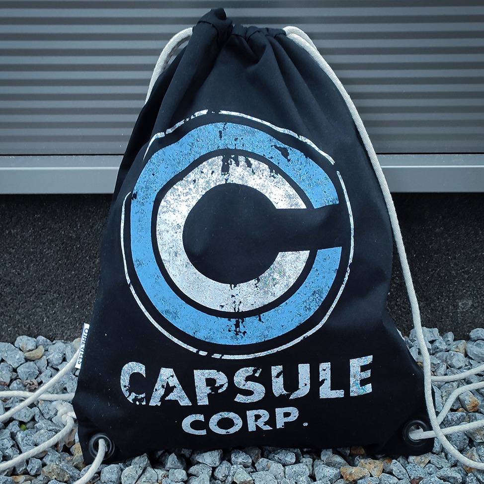 Czarny worek ze sznurkami, z nadrukiem logo Capsule Corp. w kolorze niebiesko-srebrnym, leżący na szarych kamieniach.