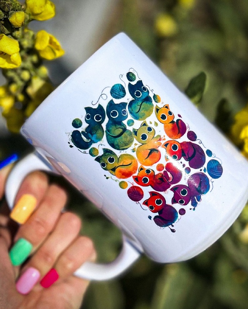 Biały kubek z kolorowym nadrukiem kotów i kłębków wełny, trzymany w dłoni z pomalowanymi na różne kolory paznokciami, na tle kwitnących żółtych kwiatów.
