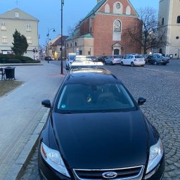 Taxi Rzeszów - Czarna taksówka Ford na brukowanym placu miejskim z zabytkowymi budynkami w tle, wczesnym rankiem. Tablica 'TAXI' na dachu pojazdu.
