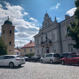 Taxi Rzeszów - Rynek z zaparkowanymi autami, budynkiem ratusza i wieżą zegarową w tle, w słoneczny dzień. Biała Toyota kombi na pierwszym planie, kostka brukowa i błękitne niebo z chmurami.