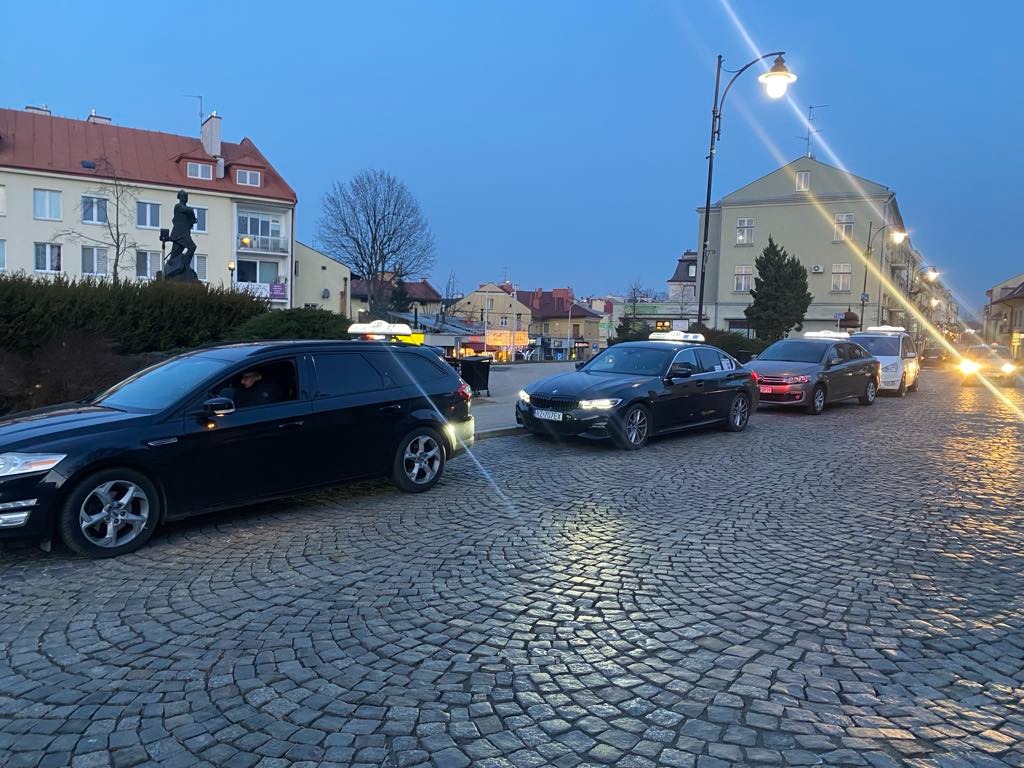 Kolejka taksówek na brukowanym placu miejskim o zmierzchu, z widocznymi latarniami i budynkami w tle.