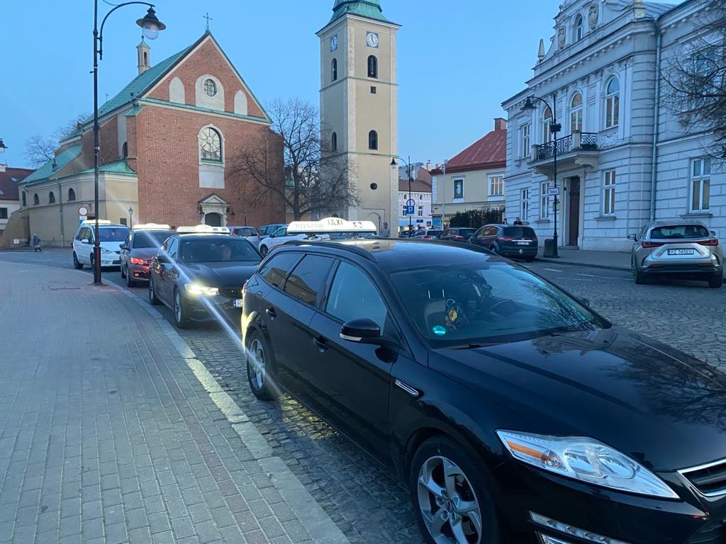 Szereg taksówek z charakterystycznymi lampami TAXI na dachu, zaparkowanych w historycznym centrum miasta z kościołem i wieżą zegarową w tle.