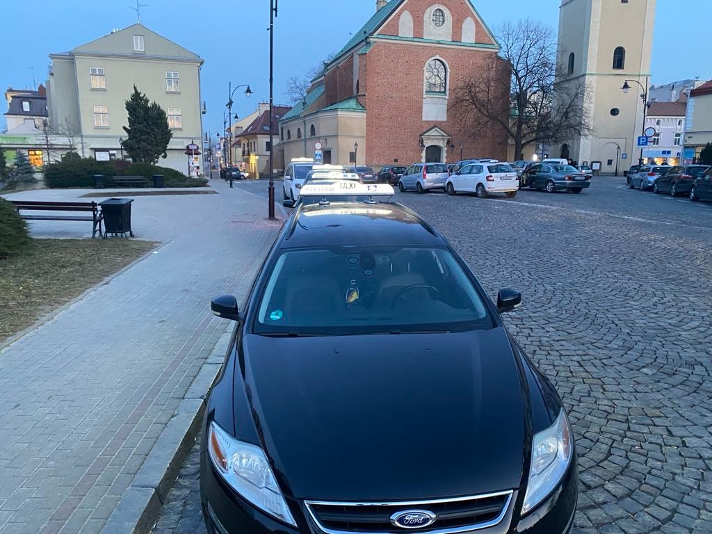 Czarna taksówka Ford na brukowanym placu miejskim z zabytkowymi budynkami w tle, wczesnym rankiem. Tablica 'TAXI' na dachu pojazdu.