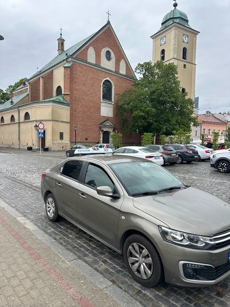 Szary samochód osobowy na tle brukowanej ulicy z kościołem i wieżą zegarową w Rzeszowie. Widoczny znak drogowy i inne zaparkowane pojazdy.