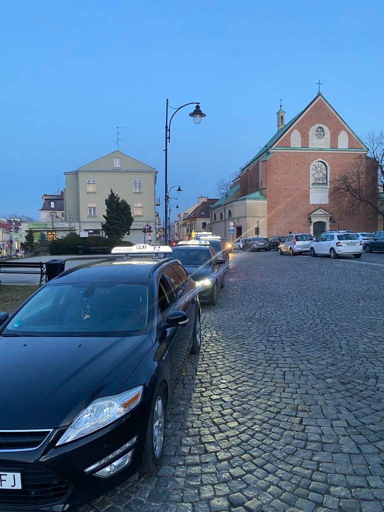 Rząd czarnych taksówek typu kombi ustawionych na brukowanej ulicy w Rzeszowie, z budynkiem kościoła i kamienicą w tle, wczesnym rankiem.