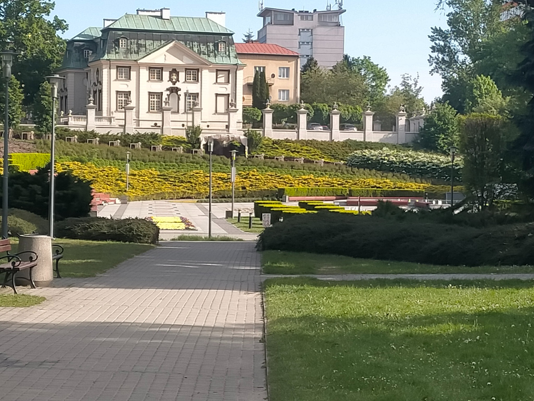 Park miejski z zadbaną zielenią, widok na klasycystyczny pałac na wzgórzu i fragment chodnika z ławkami w słoneczny dzień. Żółte kwiaty w geometrycznych rabatach.