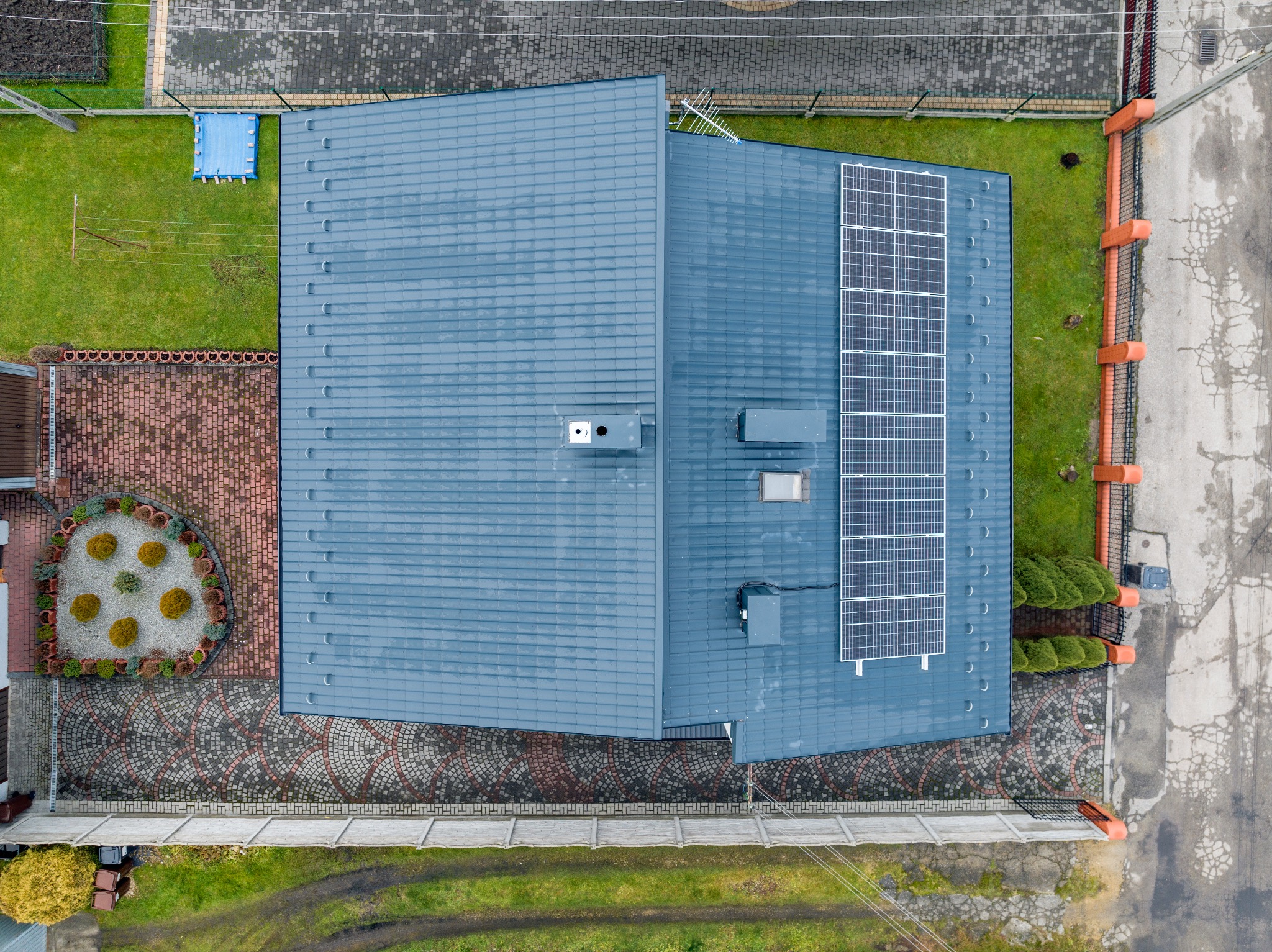Widok z góry na dom z szarym dachem pokrytym panelami słonecznymi, otoczony zielonym trawnikiem, kostką brukową i ogrodzeniem.