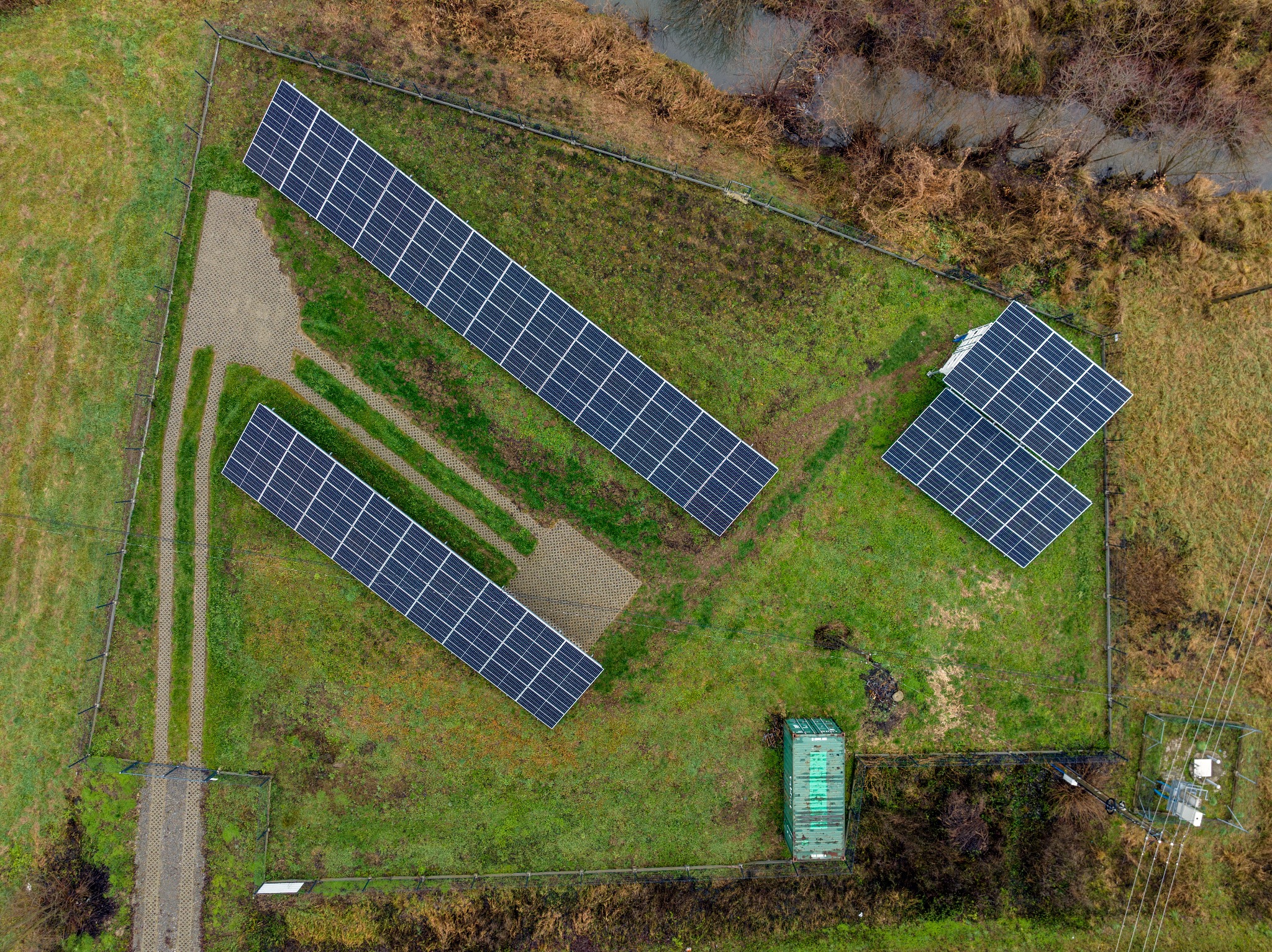Widok z góry na ogrodzoną instalację fotowoltaiczną z trzema rzędami paneli, ścieżką z ażurowych płyt i zielonym kontenerem technicznym na trawiastym terenie, obok rzeki.