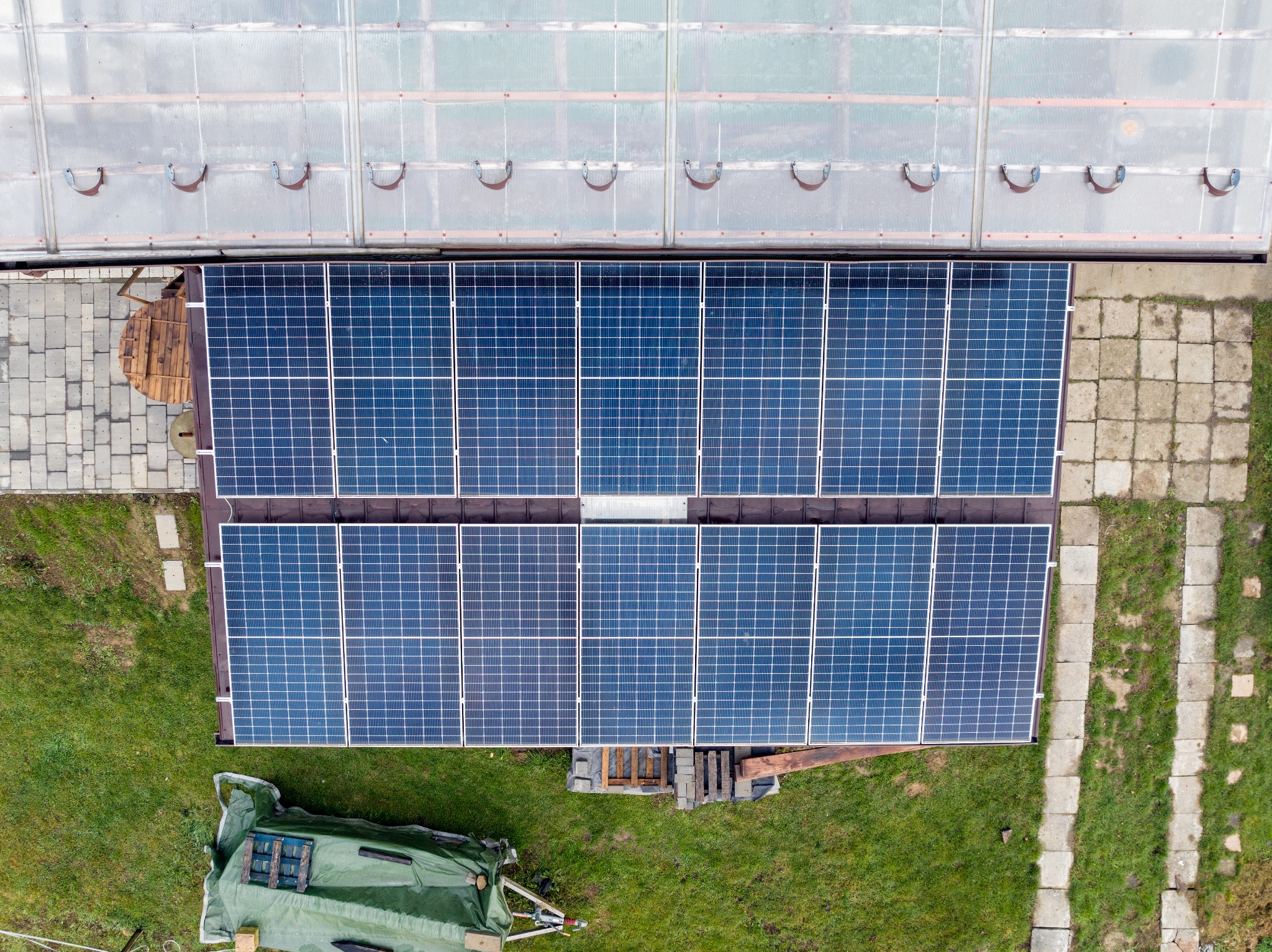 Widok z góry na panele słoneczne zamontowane na dachu budynku gospodarczego, obok szklarni, z widocznym sprzętem budowlanym pod plandeką na trawie.