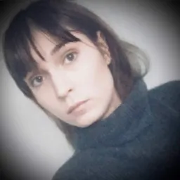 Portret kobiety z brązowymi włosami i grzywką, ubrana w ciemny golf, na rozmytym tle.