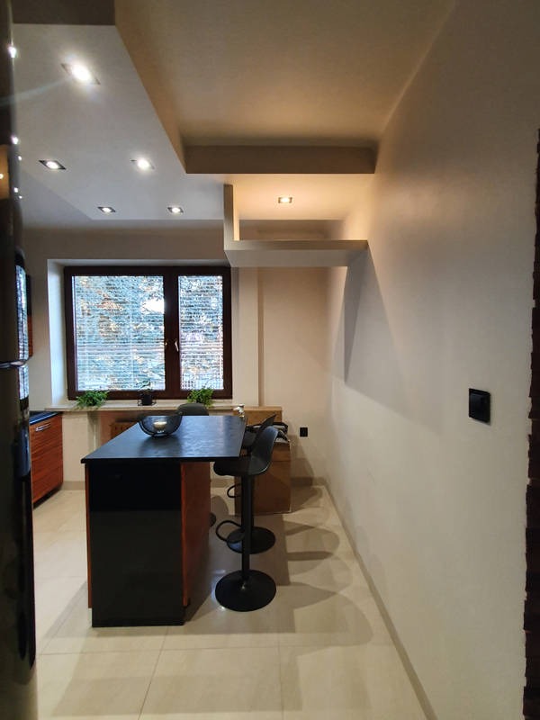 Nowoczesne wykończenie wnętrza kuchni z wyspą i hokerami, widok na okno z roletami poziomymi i roślinami na parapecie, oświetlenie punktowe w suficie podwieszanym.