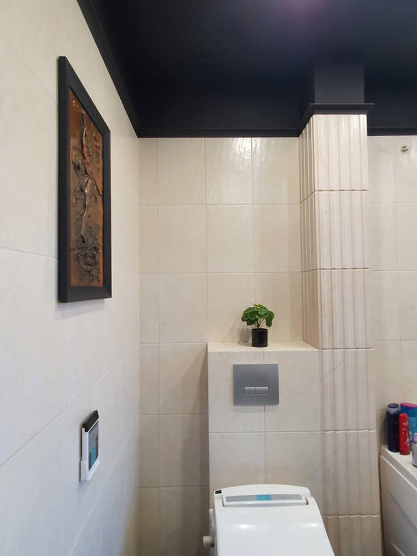 Wykończona łazienka w Bełżycach z kremowymi płytkami, czarnym sufitem podwieszanym i dekoracyjną kolumną. Na ścianie wisi obraz w ciemnej ramie, a na półce stoi roślina w doniczce.